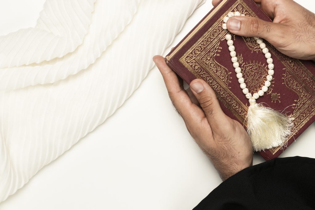 Hands holding the Holy Quran with white prayer beads, representing online Quran learning and spiritual connection.