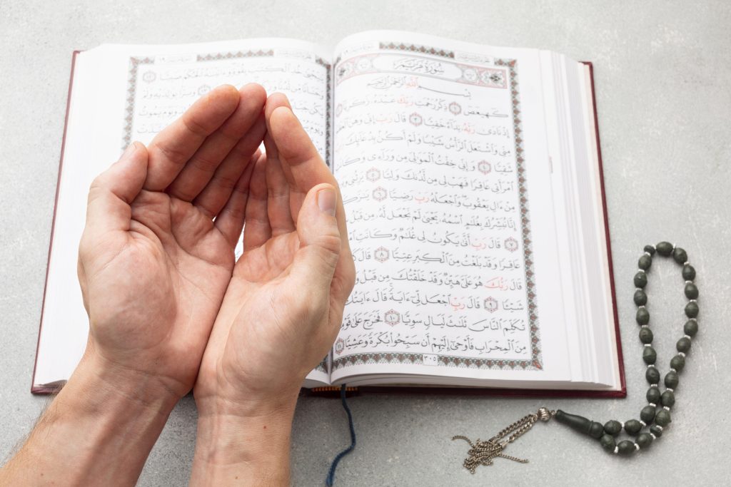 A Muslim person raising hands in prayer in front of an open Quran and prayer beads.